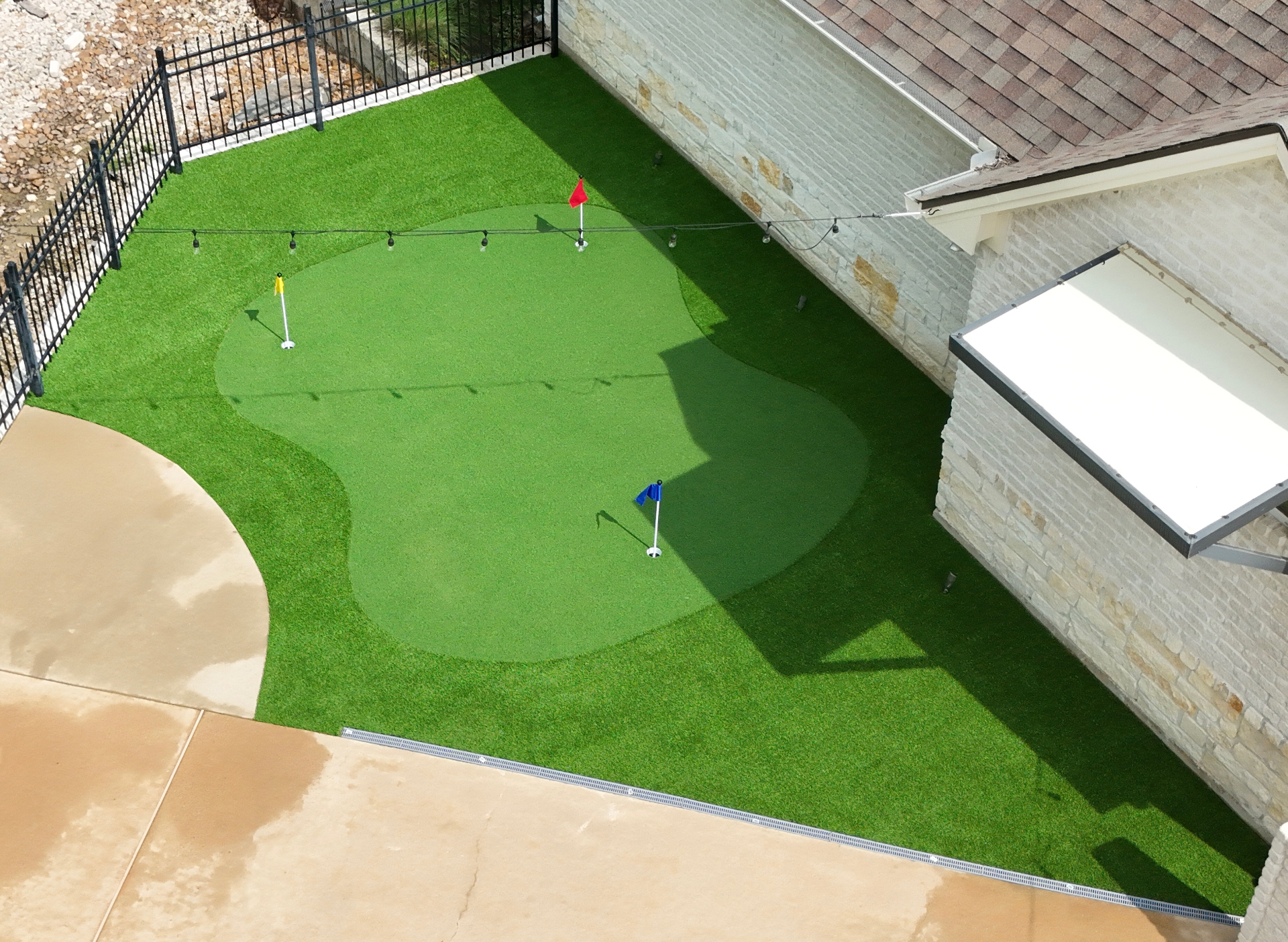Backyard putting green with red, yellow and blue flags on artificial turf next to stone home