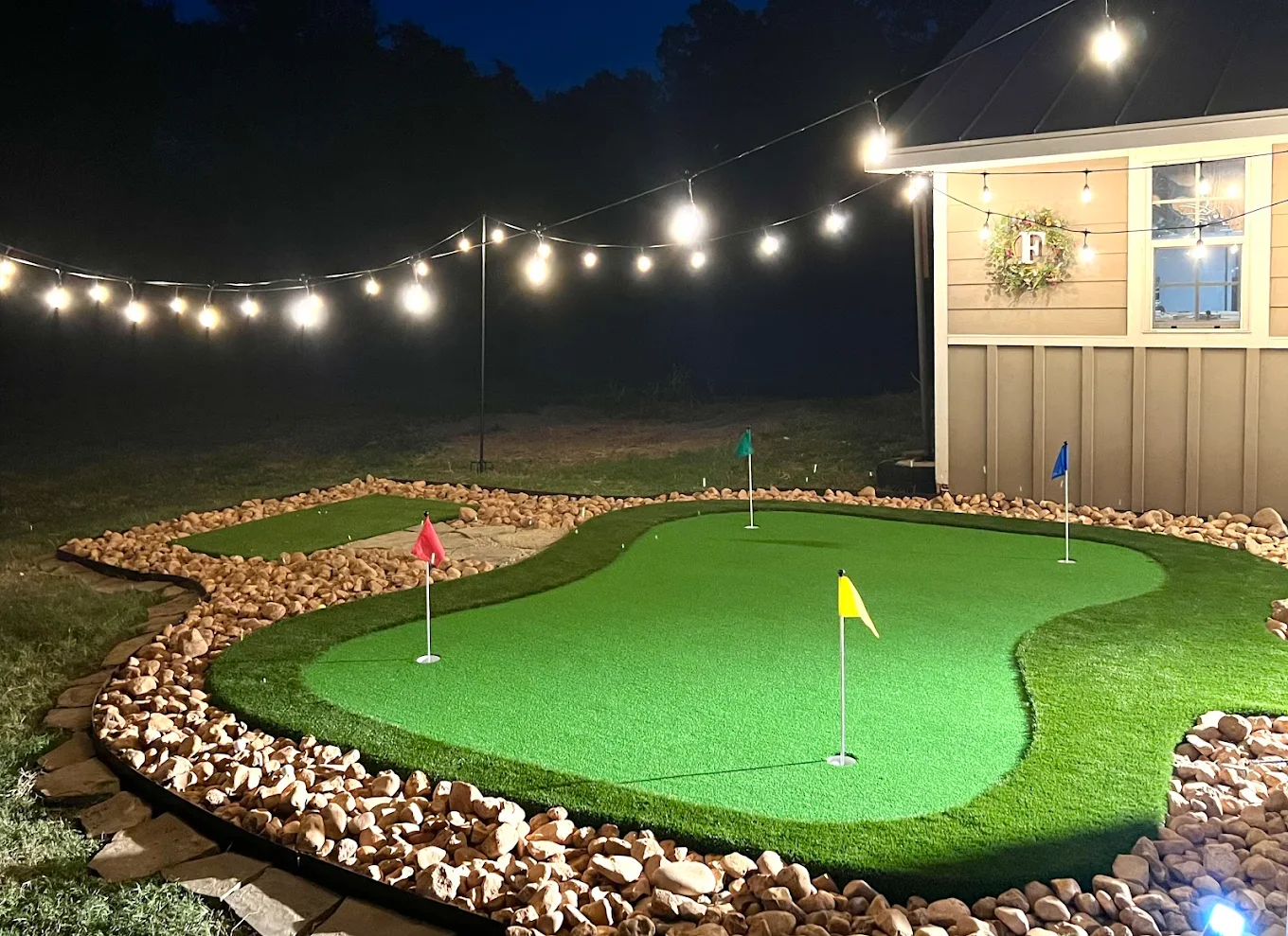 Backyard putting green at night with string lights and stone border